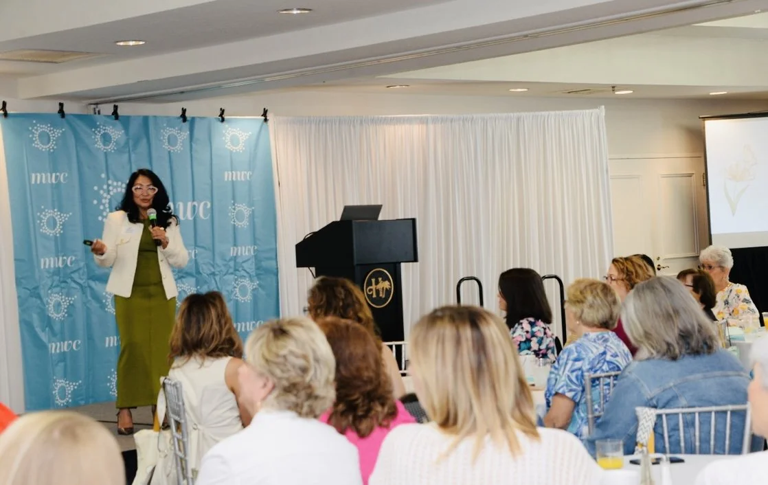 A female speaker stands on a stage holding a microphone and speaking to a room full of women sitting at tables