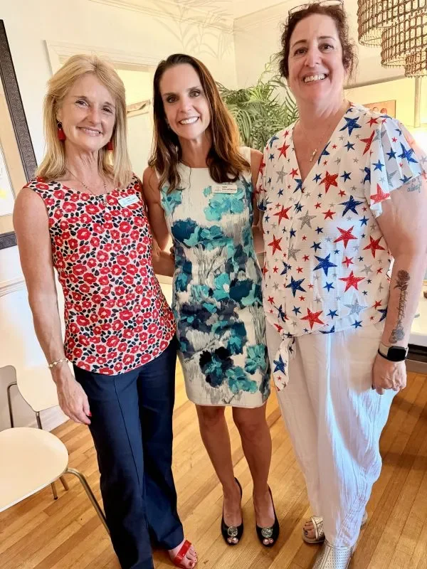 Three widows stand together with their arms wrapped around each other's backs and smiling at the camera