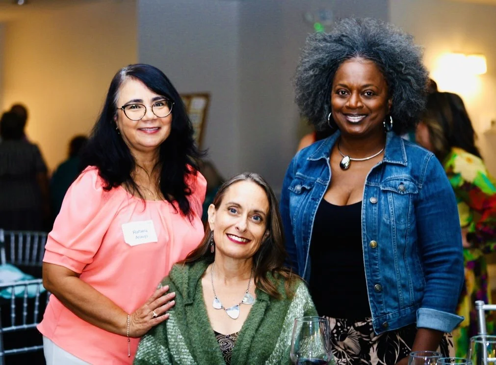 Three women pose together for a photo. The middle woman is sitting.