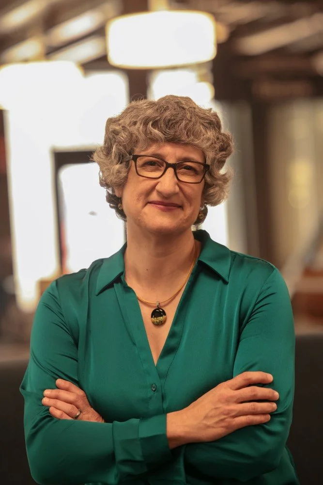 Dr. O'Connor wears an emerald green collared shirt, a large pendant necklace, black glasses, and has short cropped dark gray hair.