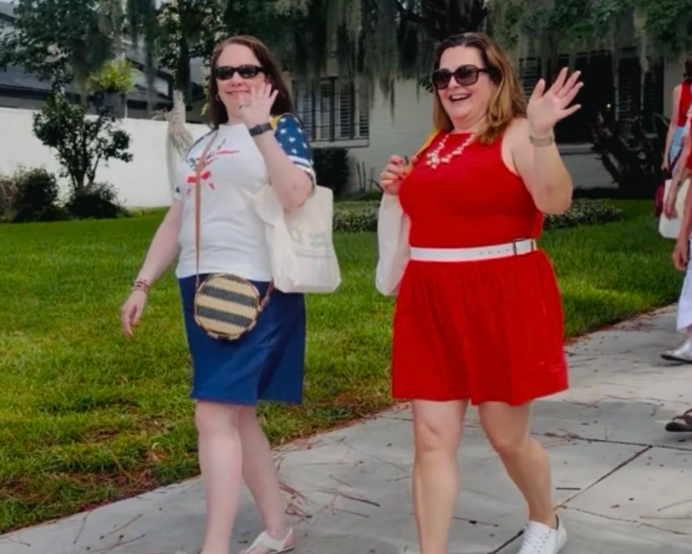 Two widows walking side by side on the sideawalk, waving at the camera. The woman on the left wears a blue skirt and a white t-shirt, and the woman on the right wears a red dress.