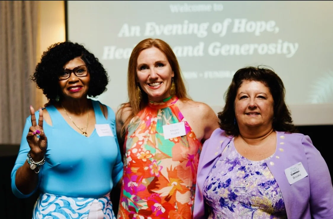 Three women stand together with their arms around each other, all looking and smiling at the camera