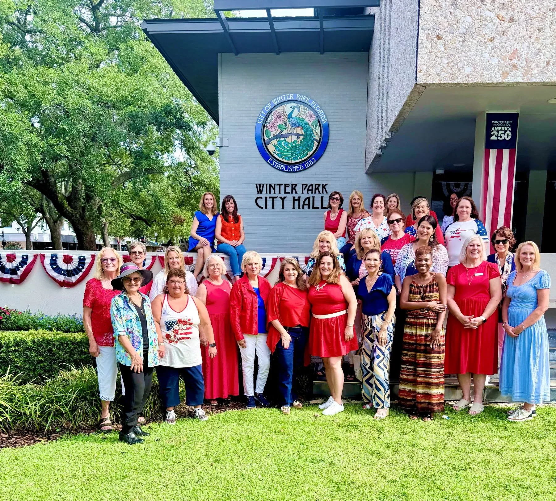 Modern Widows Club members group photo at Winter Park Florida City Hall. The women are mostly wearing red, white, and blue. About 5 sit on a cement wall behind a group of about 20 women standing