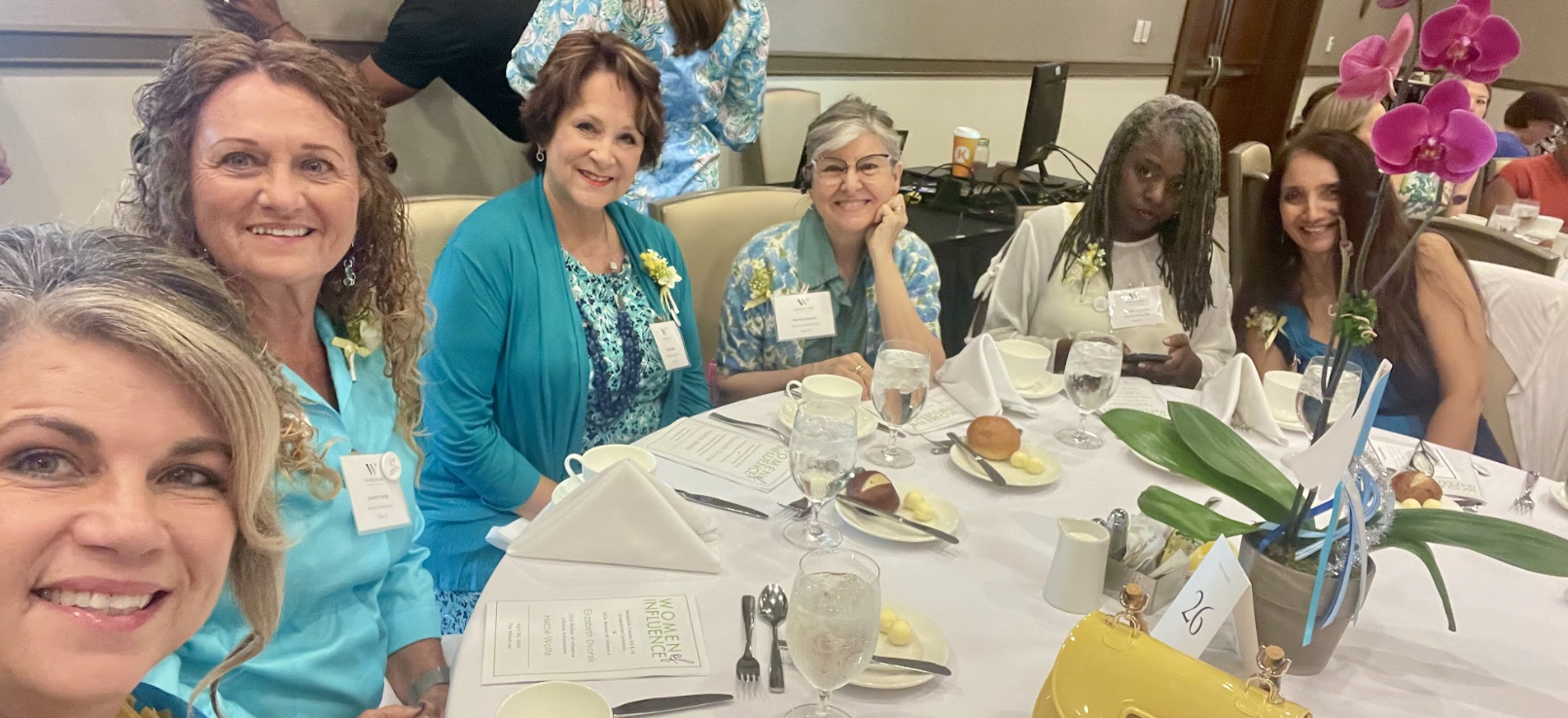 A group of six women sit together at a round table, all looking at and smiling at the camera for a selfie. They are mostly wearing shades of turquoise and teal and are at a Modern Widows Club event.