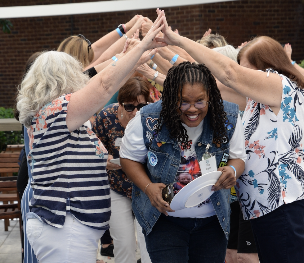 A Black woman walks underneath a bridge made up of other women's arms leaning together in a triangle
