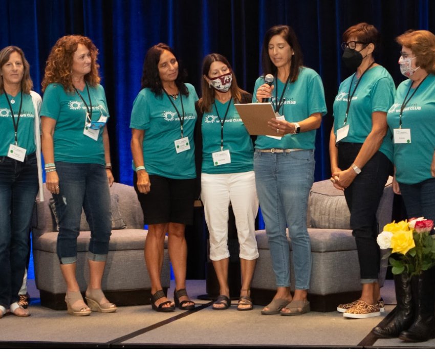 Group of seven women standing together, with one speaking into a microphone