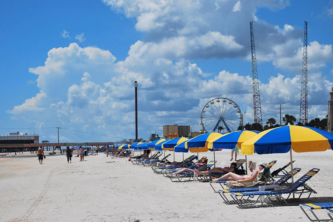 Sandy beach with a straight line of lounge chairs and blue and yellow beach umbrellas and a ferris wheel in the background