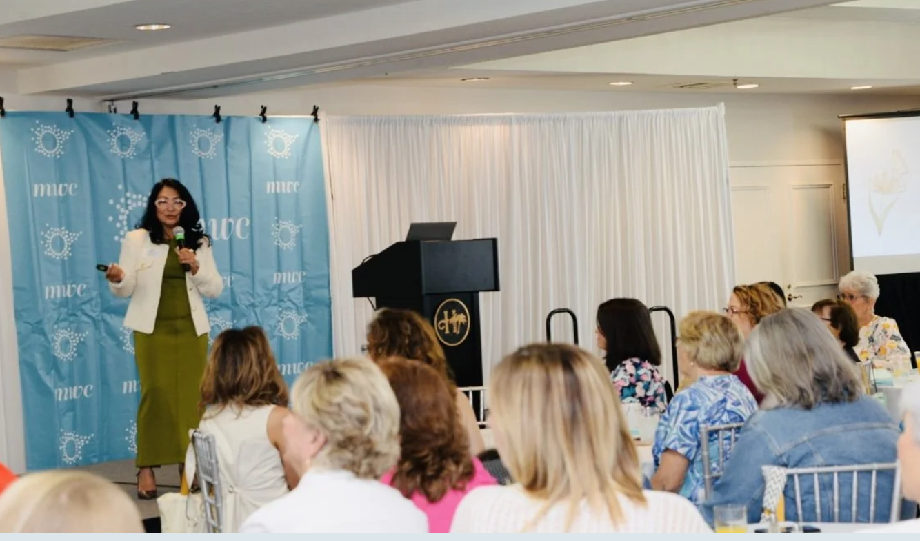 A woman giving a presentation on stage with a blue backdrop and audience watching in a conference room.