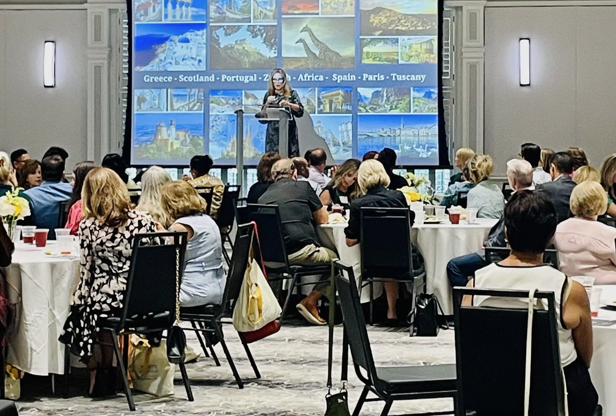 Woman speaking in front of a crowded room of men and women sitting at round banquet tables. There is a slide on the screen behind her with the words Greece, Scotland, Portugal, Africa, Spain, Paris, Tuscany.