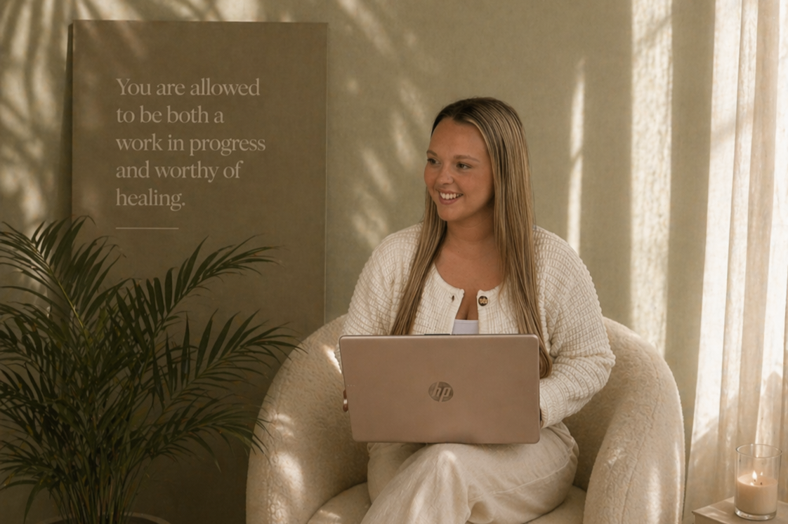 A woman with long blonde hair sitting on a cream-colored armchair, smiling while holding a silver HP laptop. There is a large green plant on her left and a candle on a small table to her right. Behind her, a sign on the wall reads, 'You are allowed to be both a work in progress and worthy of healing.' Sunlight filters through beige curtains.