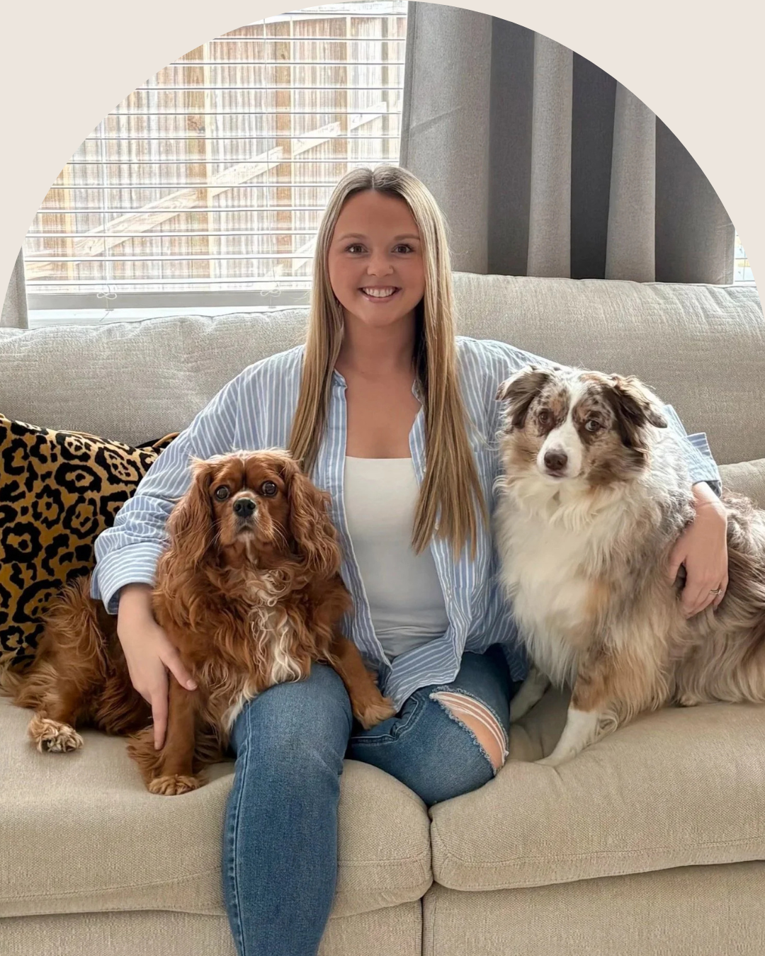 A woman sitting on a beige sofa with a Cavalier King Charles Spaniel on her left and an Australian Shepherd on her right, smiling in a well-lit living room with a window and grey curtains behind her.