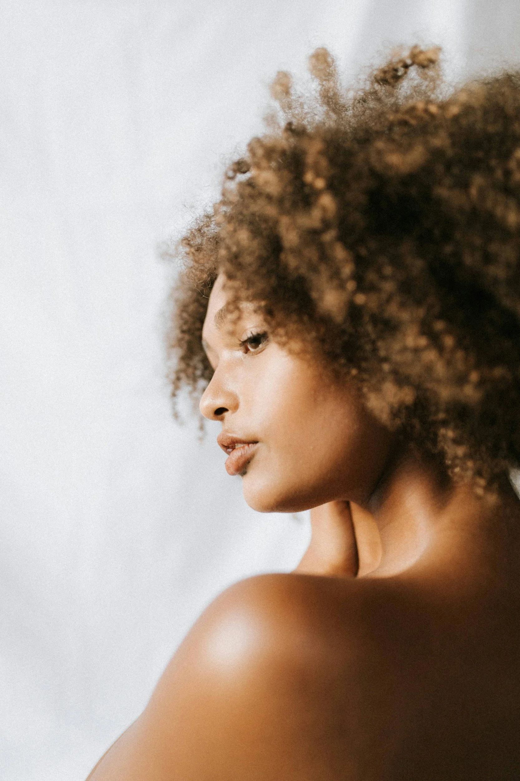 Side profile of a woman with curly hair, looking downward with a neutral expression, bare shoulders, and smooth skin, against a plain light background.