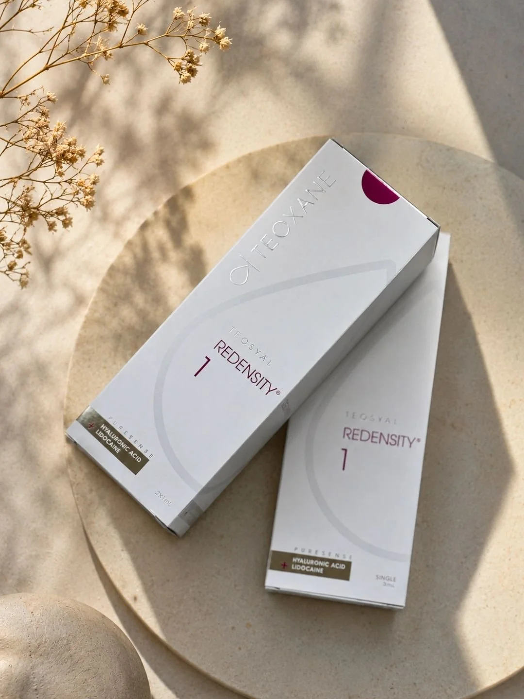 Boxes of Teoxane Redensity 1 hyaluronic acid and lidocaine dermal filler on a table with dried flowers and shadows.