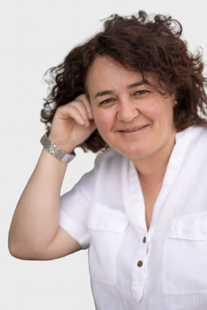 Retrato de una mujer de cabello rizado y oscuro, sonriendo y descansando la cabeza en su mano, usando blusa blanca, en un ambiente exterior.