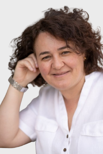 Retrato de una mujer de cabello rizado y oscuro, sonriendo y descansando la cabeza en su mano, usando blusa blanca, en un ambiente exterior.