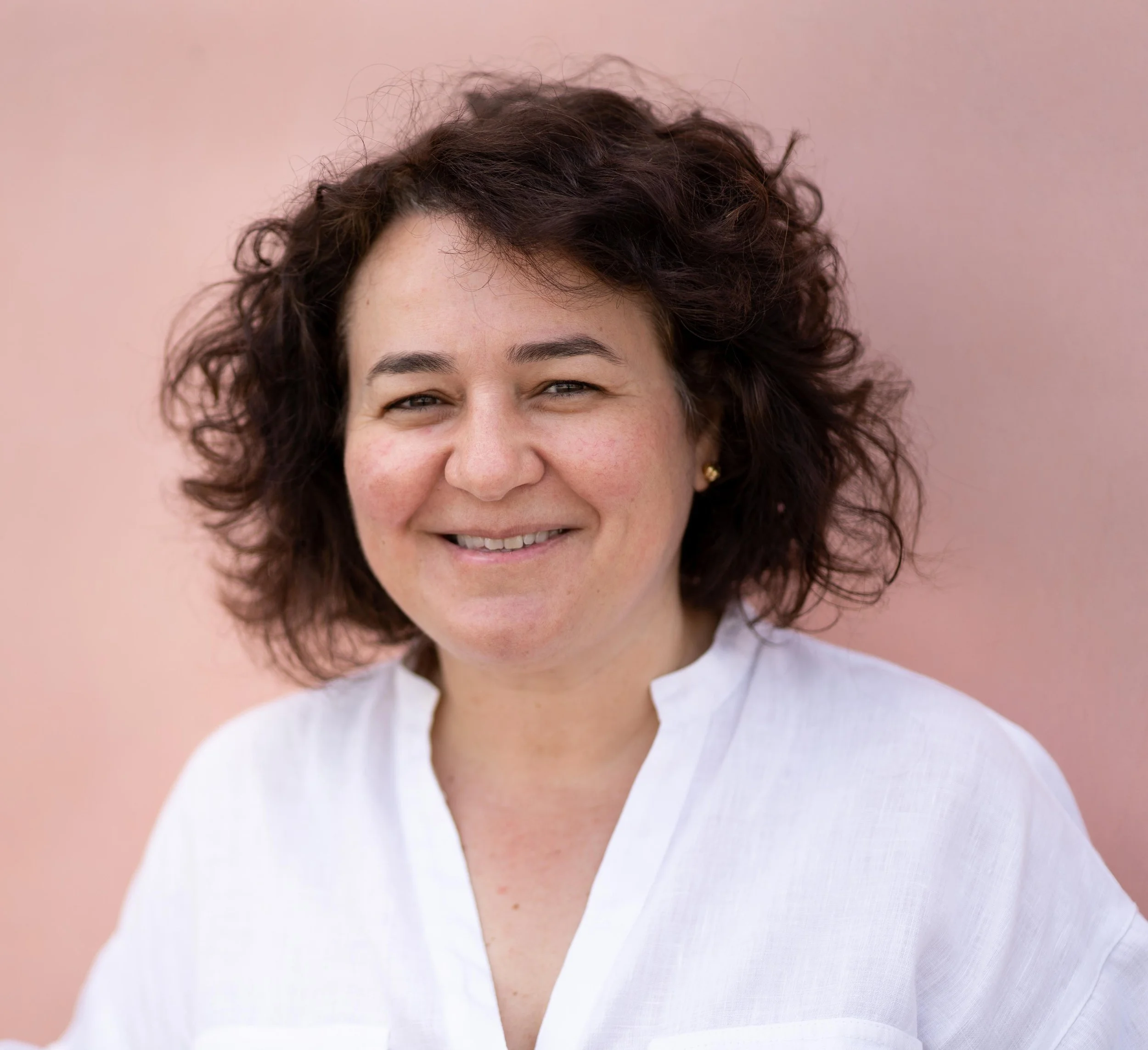 Retrato de una mujer de cabello rizado y oscuro, sonriendo y descansando la cabeza en su mano, usando blusa blanca, en un ambiente exterior.