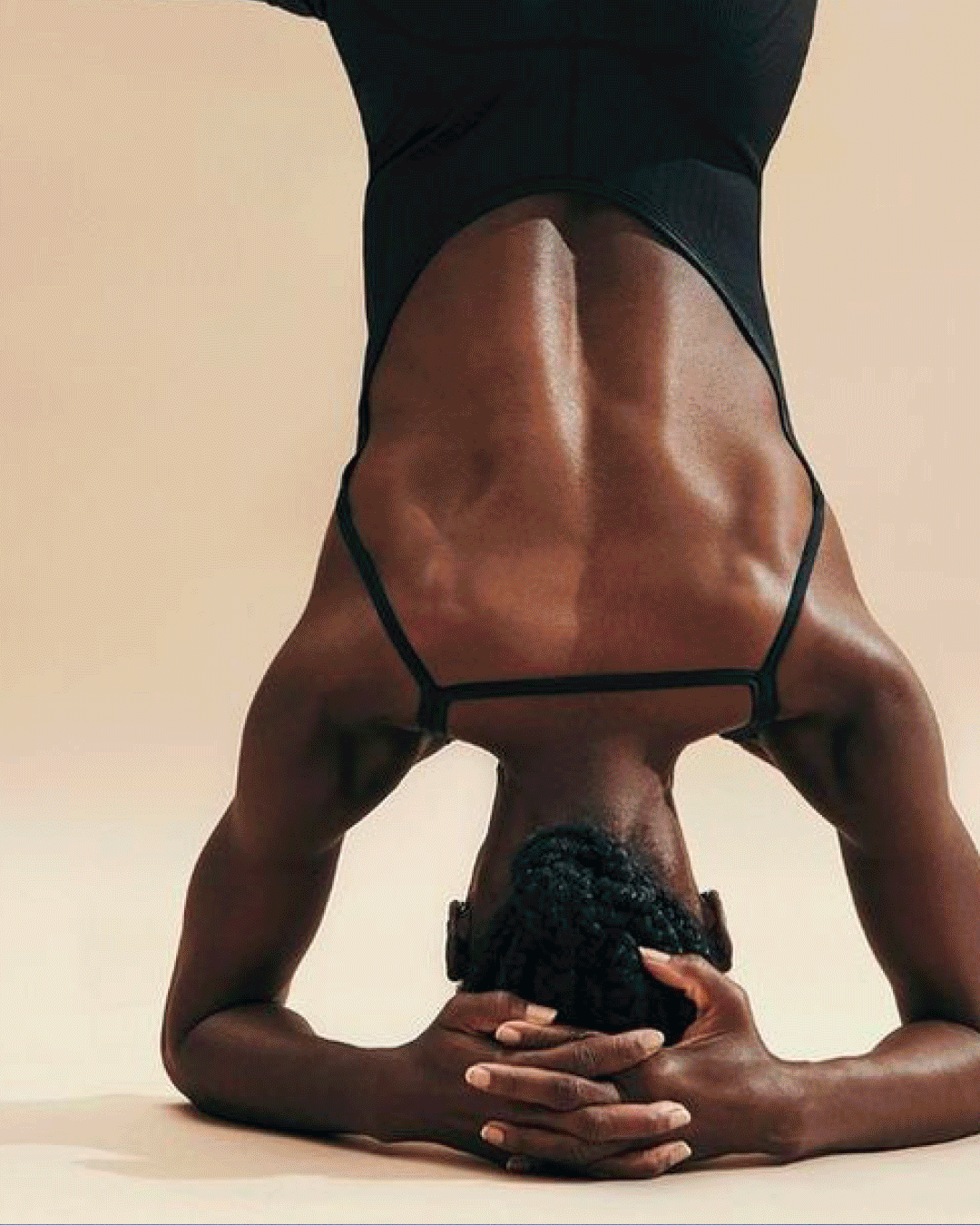 Une femme noire effectuant une posture de yoga, en position de tête-à-terre avec les bras croisés sous la tête, portant une tenue de sport noire.