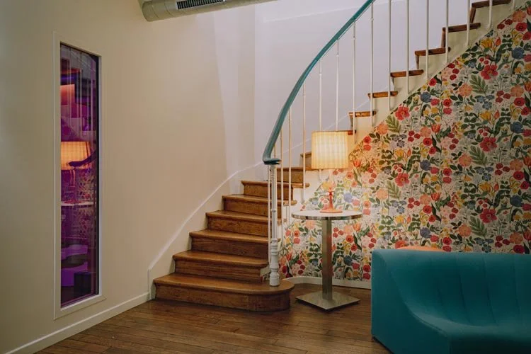 Escalier en bois avec une rampe bleue, mur décoré de papier peint floral, coin avec un petit plateau rond, lampe de table, et canapé bleu turquoise, dans une pièce intérieure