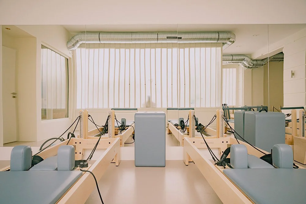 Salle de physiothérapie avec plusieurs tables de traitement équipées de coussins et appareils de physiothérapie, grands fenêtres avec stores et conduits de ventilation visibles au plafond.