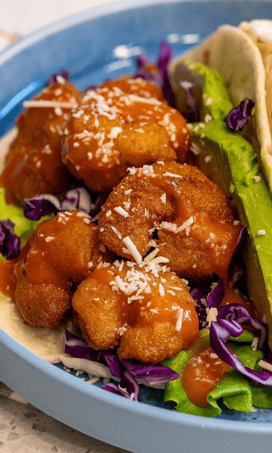 Close-up of a bowl of fried chicken pieces with sauce and shredded cheese, served with shredded purple cabbage, green lettuce, and a taco shell at the side.