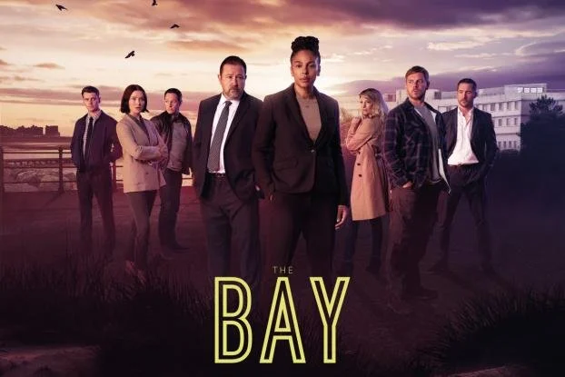 Group of seven diverse people standing outdoors during sunset with city buildings in the background, promotional poster for the TV show 'The Bay'.