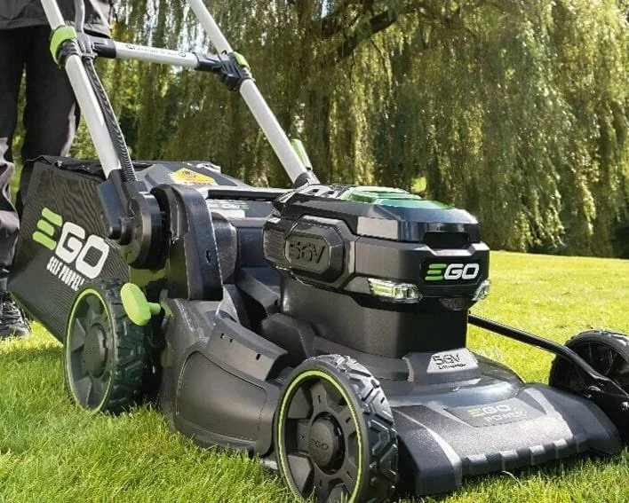 A close-up view of a black and green cordless electric lawn mower on a grassy lawn with trees in the background.