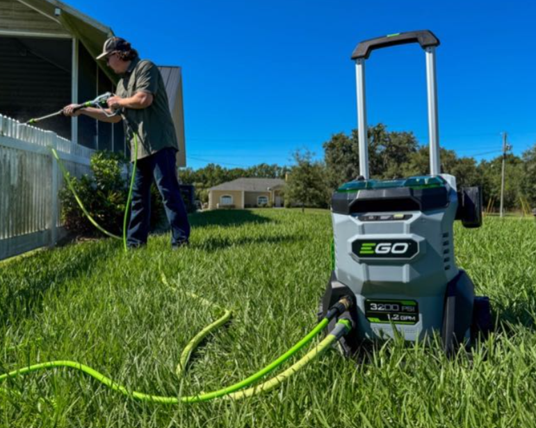 A man using a pressure washer on a house's exterior wall in a backyard with a green lawn, with a portable pressure washer machine in the foreground.