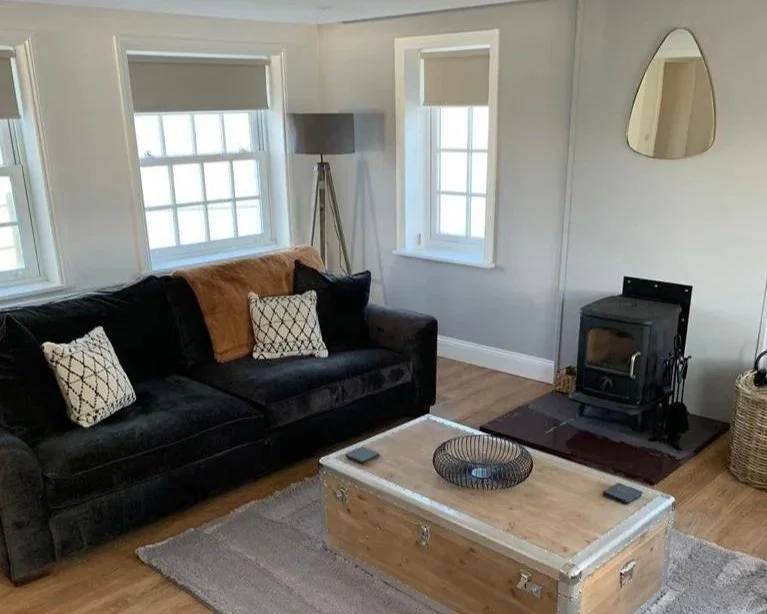 Living room with a black couch, decorative pillows, a wooden trunk as a coffee table, a wood stove, a floor lamp, and windows with natural light.