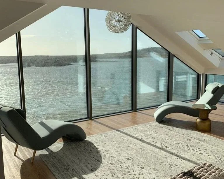 Modern living room with large floor-to-ceiling windows overlooking a body of water, two gray lounge chairs, a light-colored area rug, and a round pendant light hanging from the ceiling.
