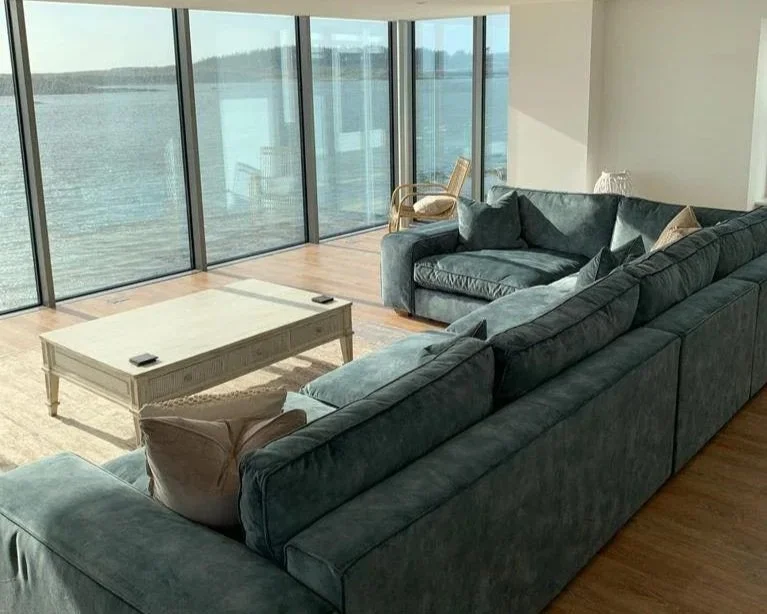 Living room with large L-shaped blue sofa, cream coffee table, wicker chair, and view of water through floor-to-ceiling windows