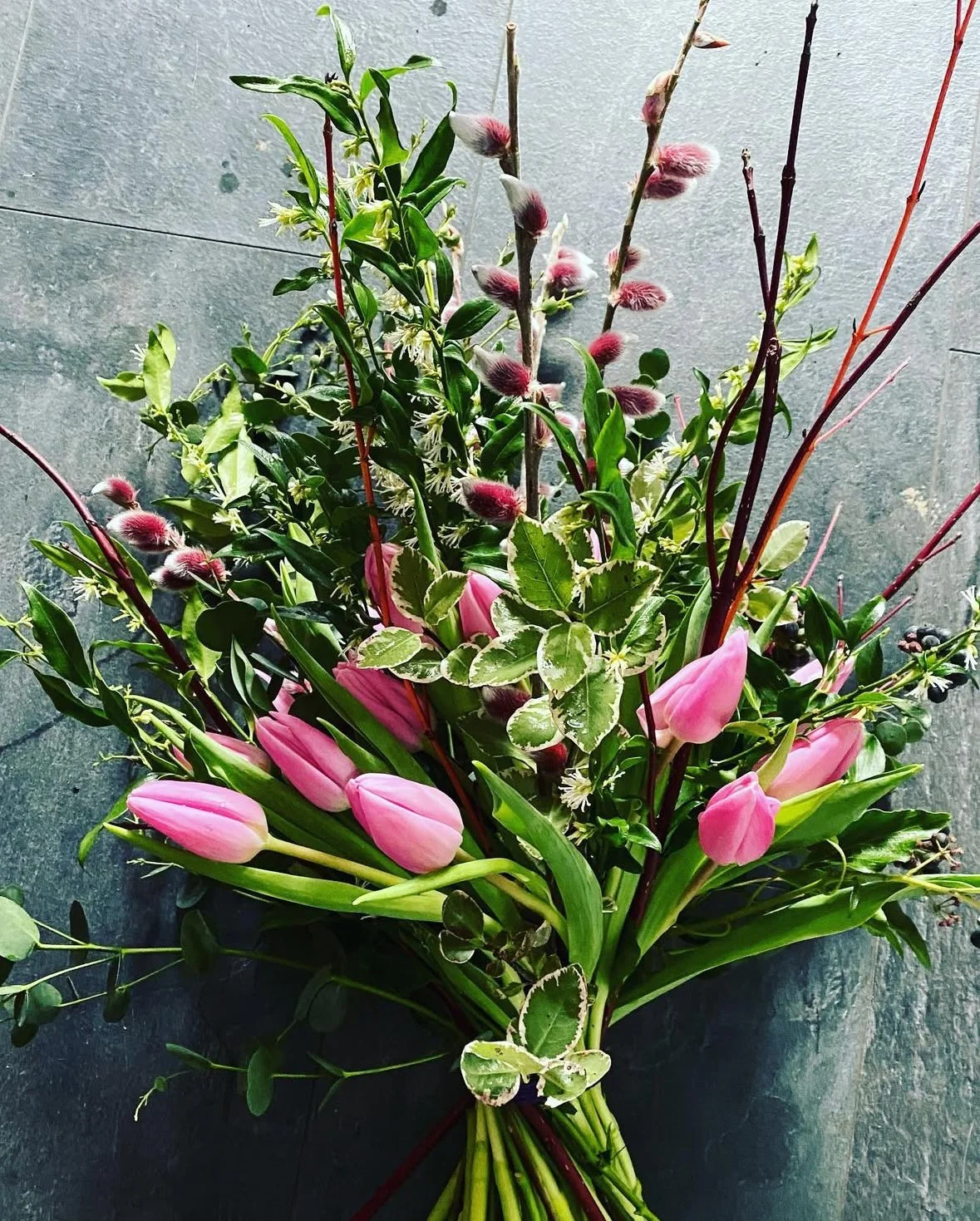 A bouquet of pink tulips, variegated leaves, and pussy willow branches arranged on a gray surface.
