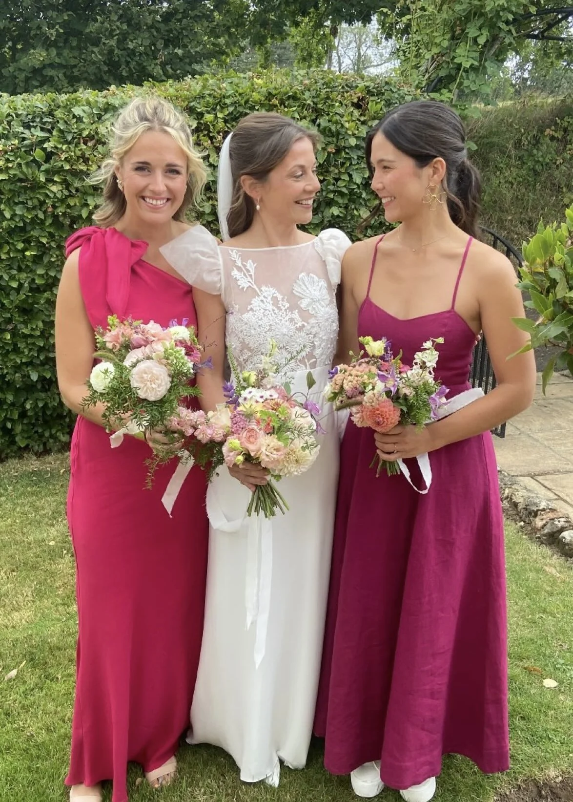 Three women in wedding attire holding bouquets of flowers outside in a garden setting, smiling and looking at each other.