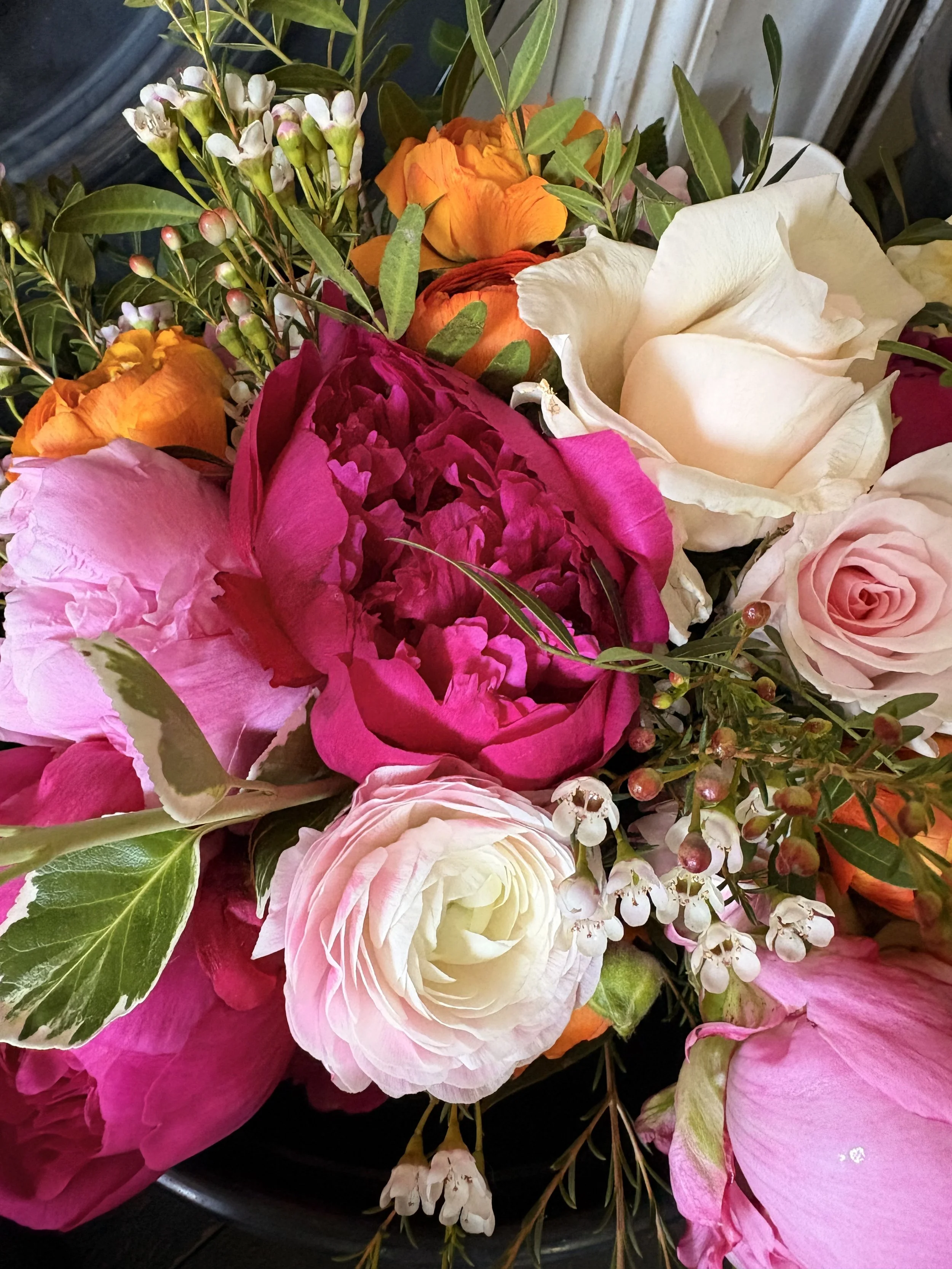 A colorful bouquet of various flowers including pink and white roses, pink peonies, orange and yellow tulips, and small white flowers with green leaves.