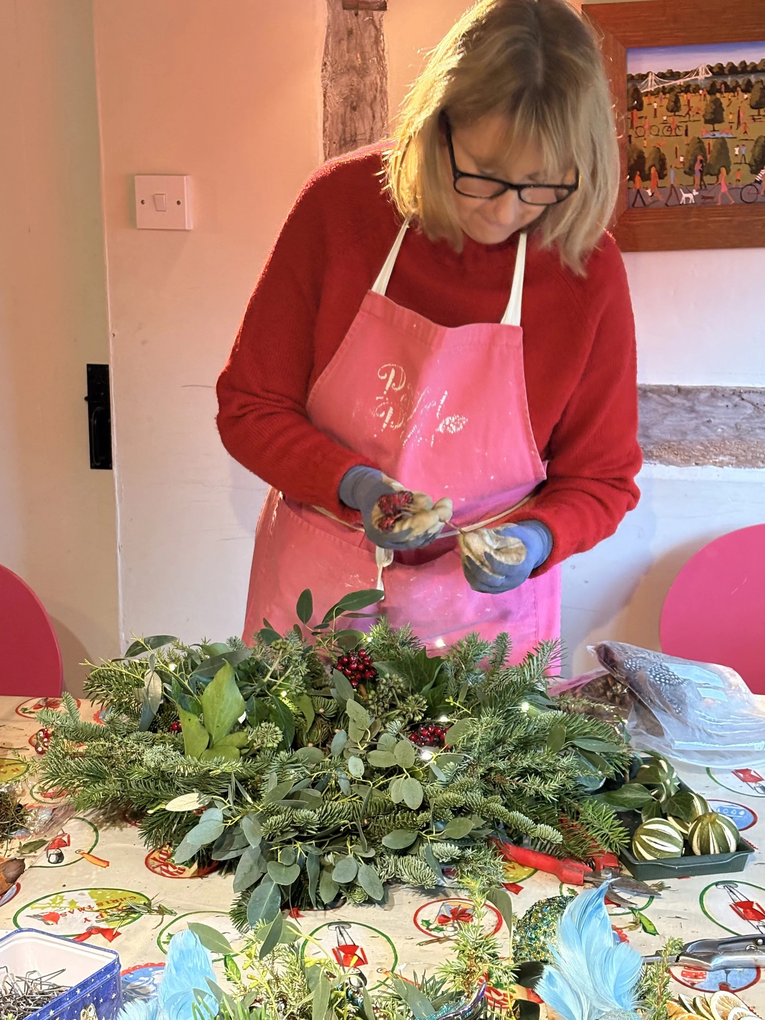 A woman wearing glasses, a red sweater, pink apron, and gloves is creating a Christmas centerpiece with greenery, berries, and decorative elements on a festive table.