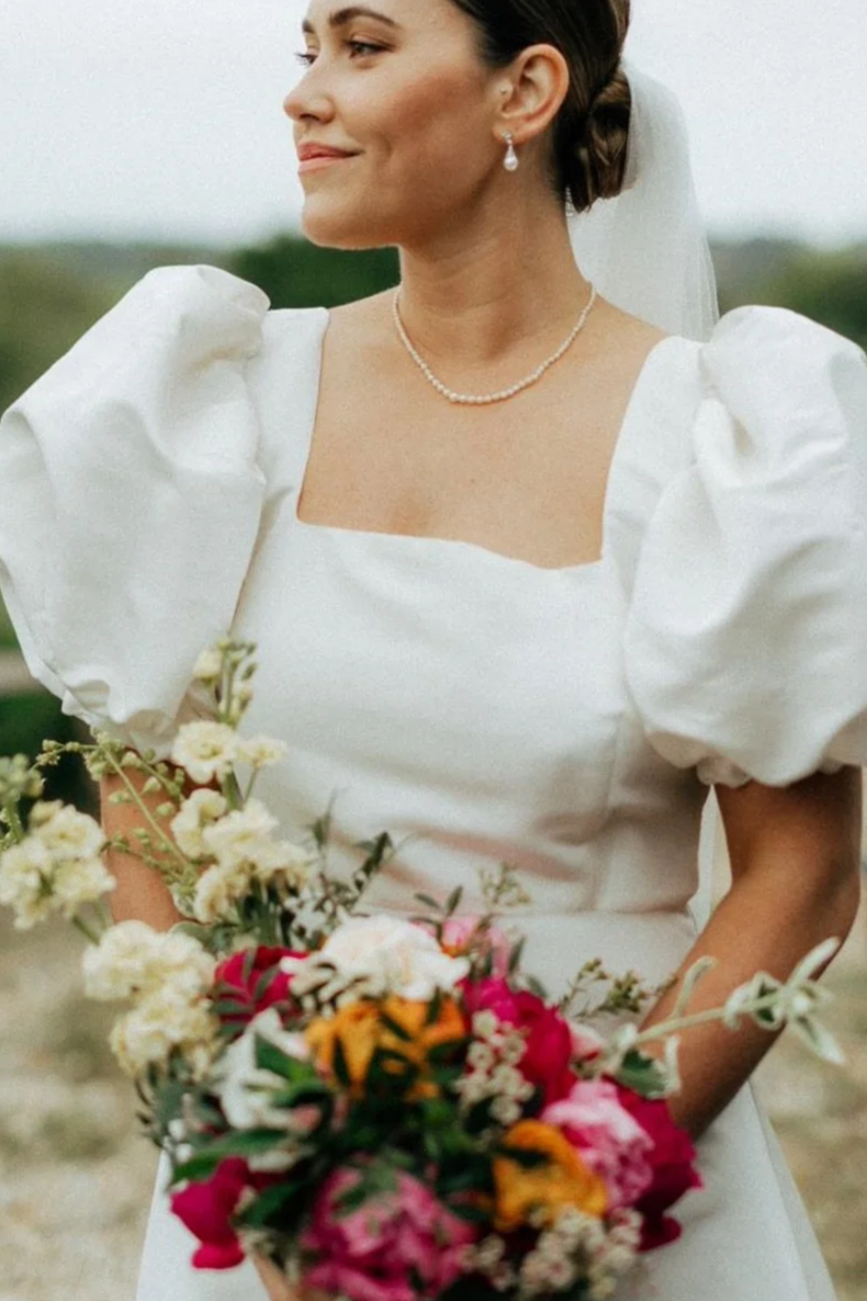A woman in a white wedding dress with puffed sleeves holding a colorful bouquet of flowers. She is wearing pearl jewelry and has her hair styled in an elegant updo with a veil. The background is blurred, indicating an outdoor setting.