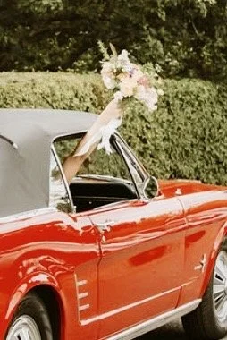 A person holding a bouquet of flowers outside a red vintage convertible car.