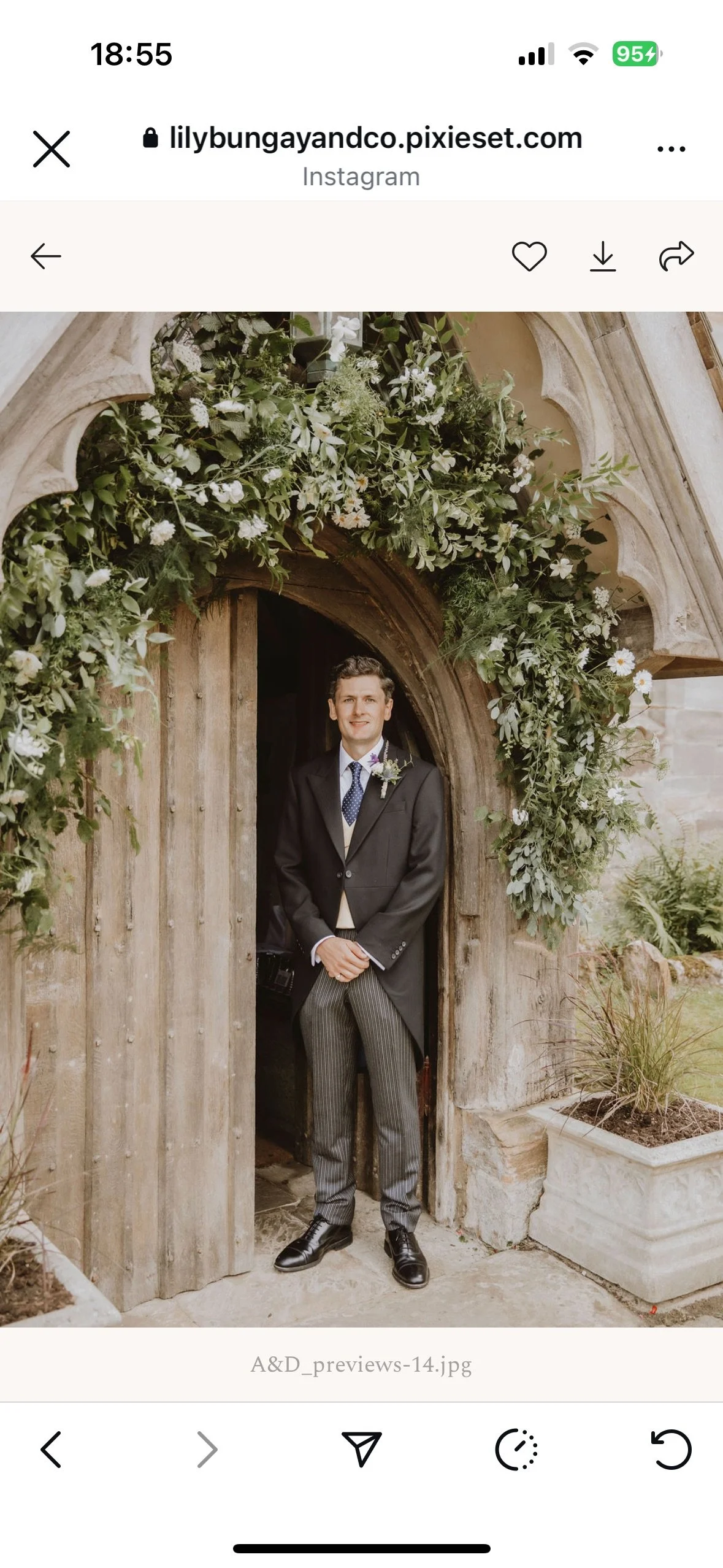 A young man dressed in a formal tuxedo and striped pants standing in a wooden doorway decorated with lush greenery and white flowers, likely at a wedding or special event.