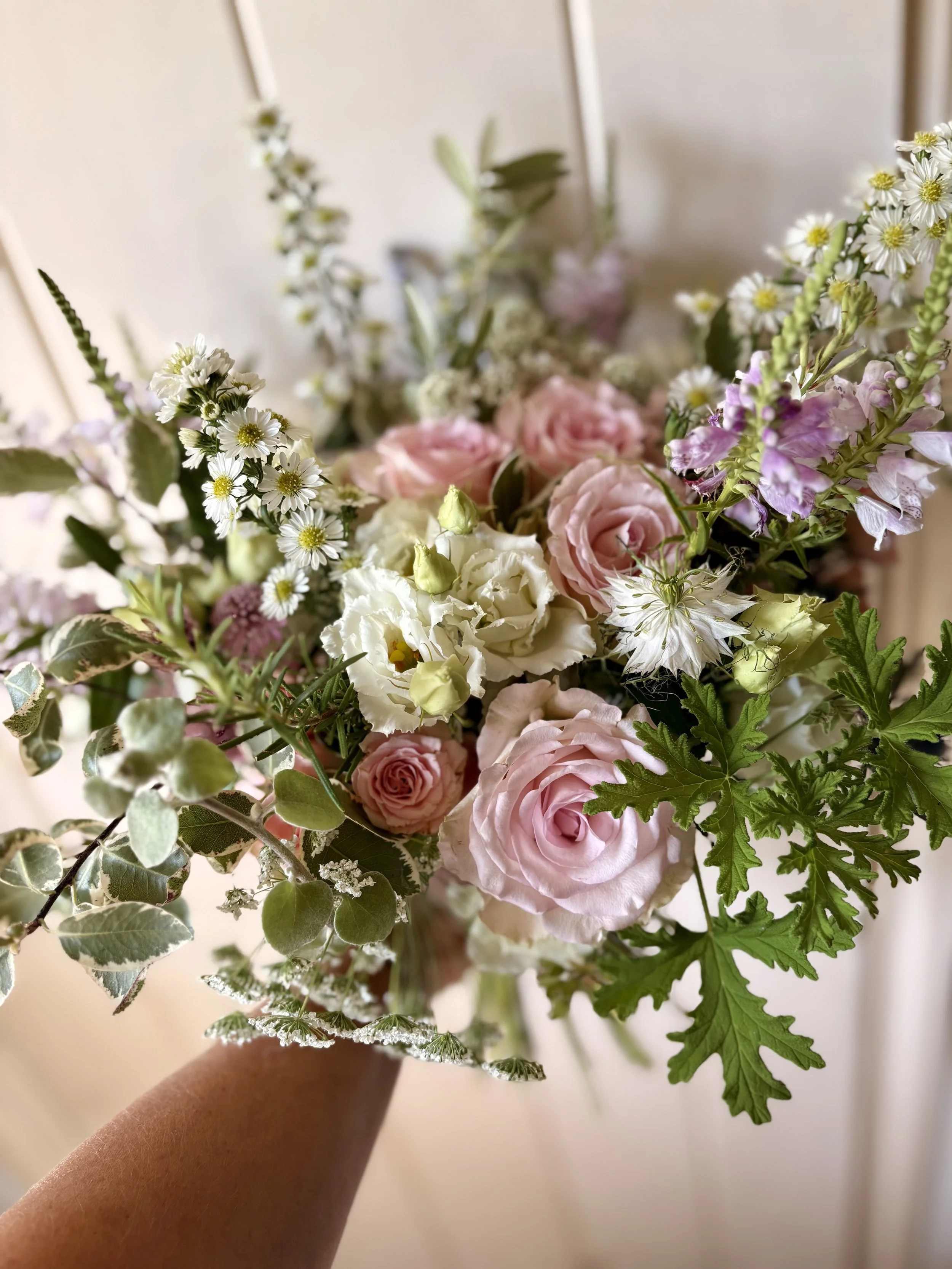 A close-up of a colorful bouquet of pink, white, and purple flowers with greenery, held in a person's hand.