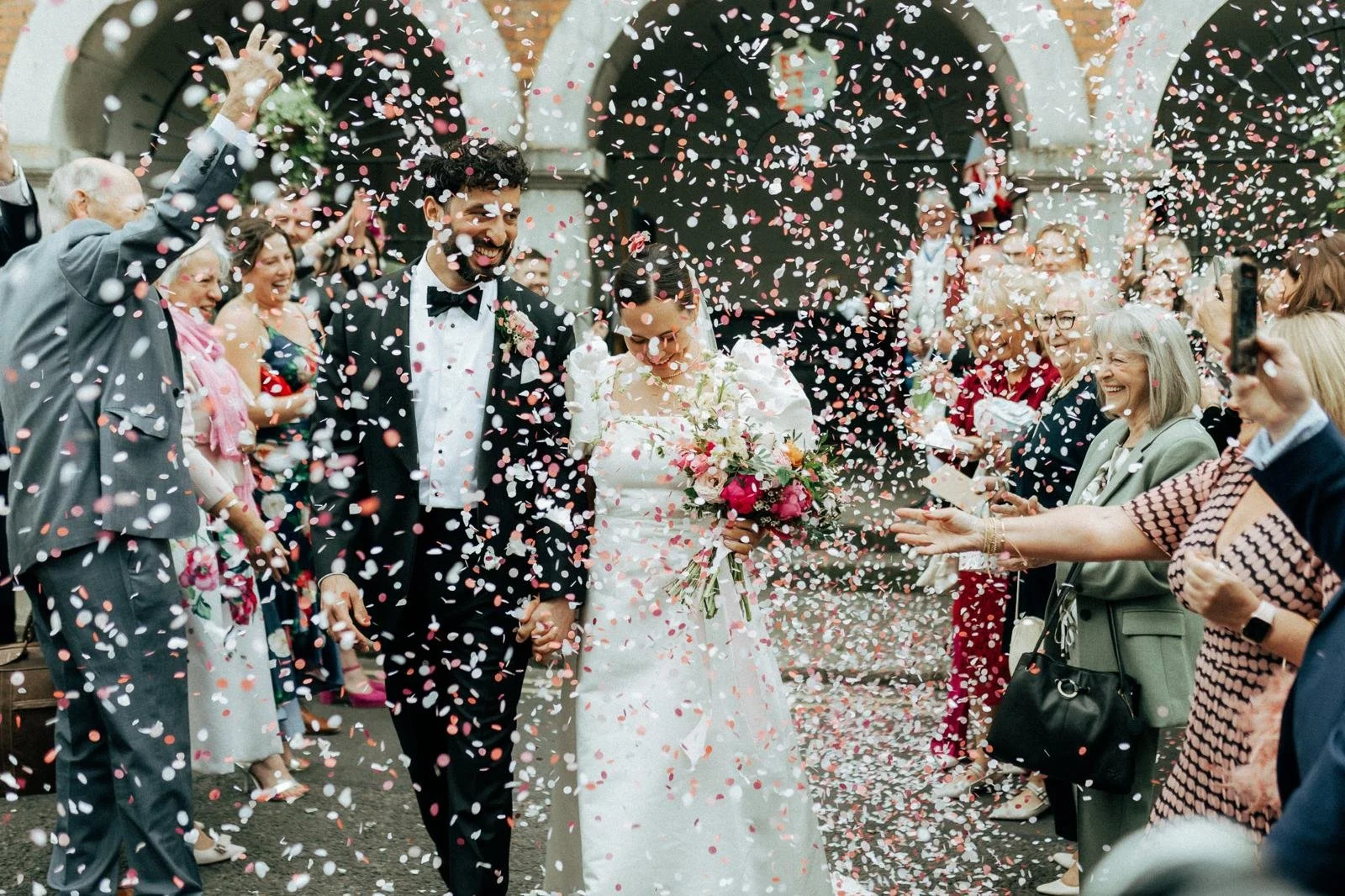 A newlywed couple walking through a shower of pink and white confetti outside a building, surrounded by smiling guests.