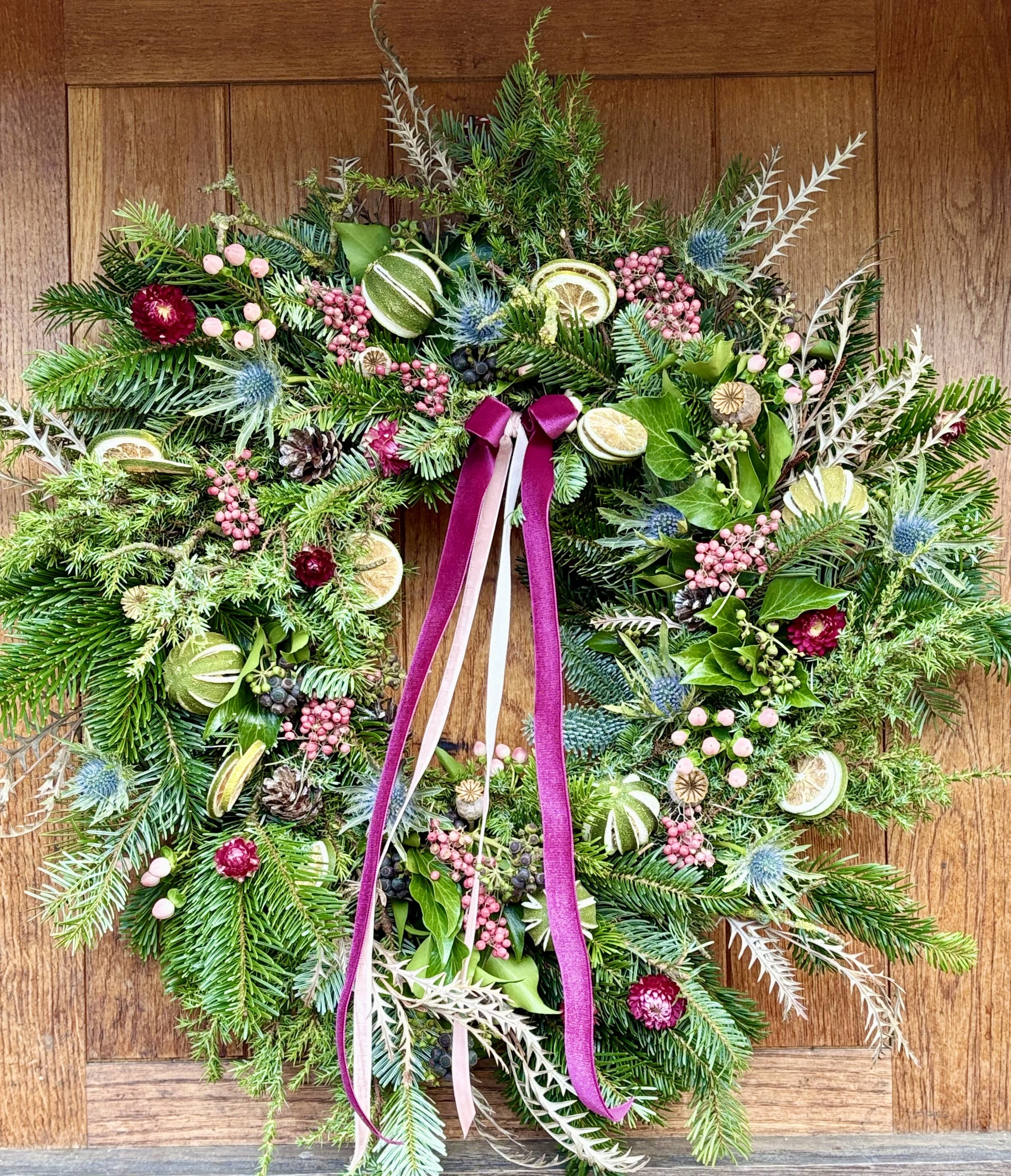 A decorative Christmas wreath hanging on a wooden door, adorned with various green foliage, dried citrus slices, pinecones, berries, blue thistle flowers, and a purple ribbon in the center.
