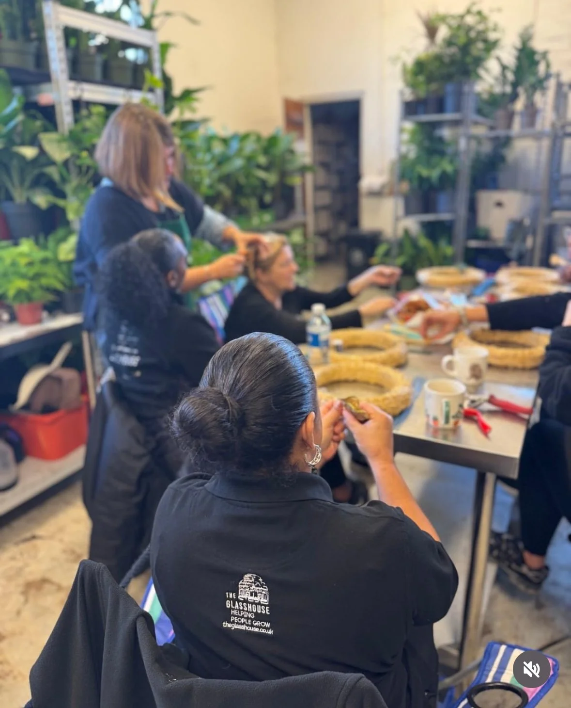 A group of women participating in a wreath-making workshop in a plant-filled indoor space.