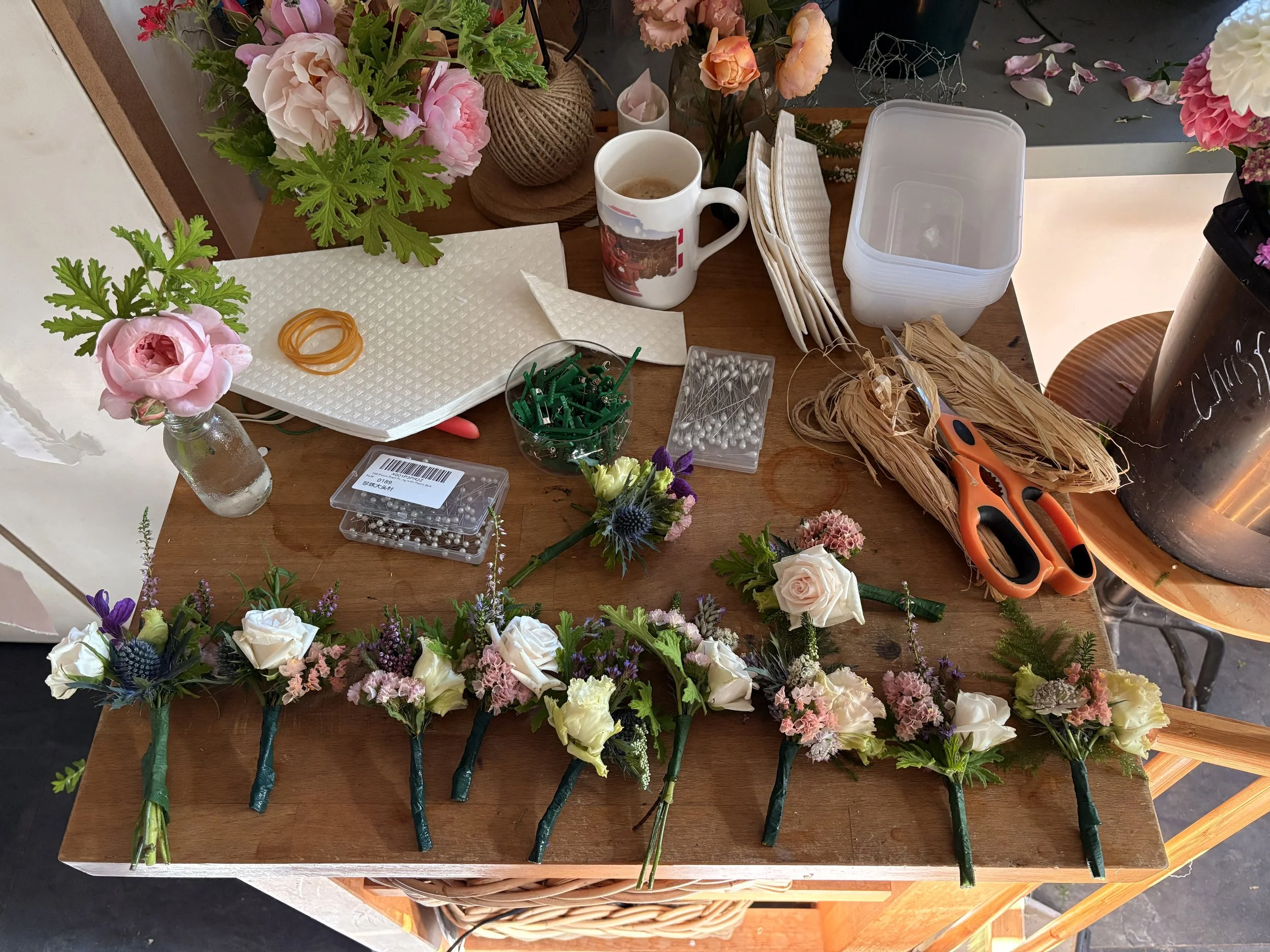 A wooden work table with floral arrangements, scissors, pins, rubber bands, a mug, and other flower arranging tools, surrounded by flowers and supplies.
