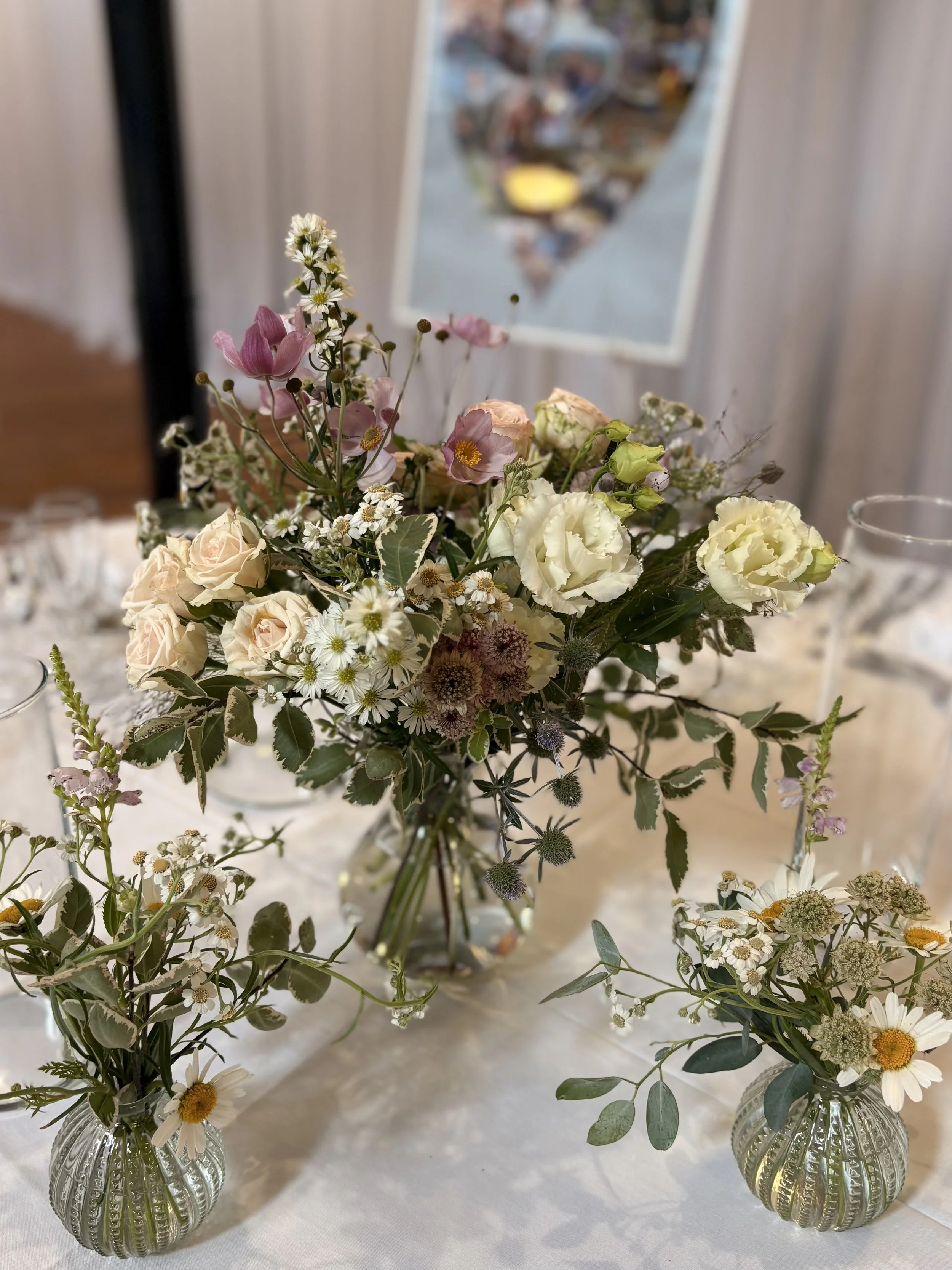 A floral centerpiece with pink, white, and purple flowers on a table, with two smaller vases of daisies and greenery surrounding it.