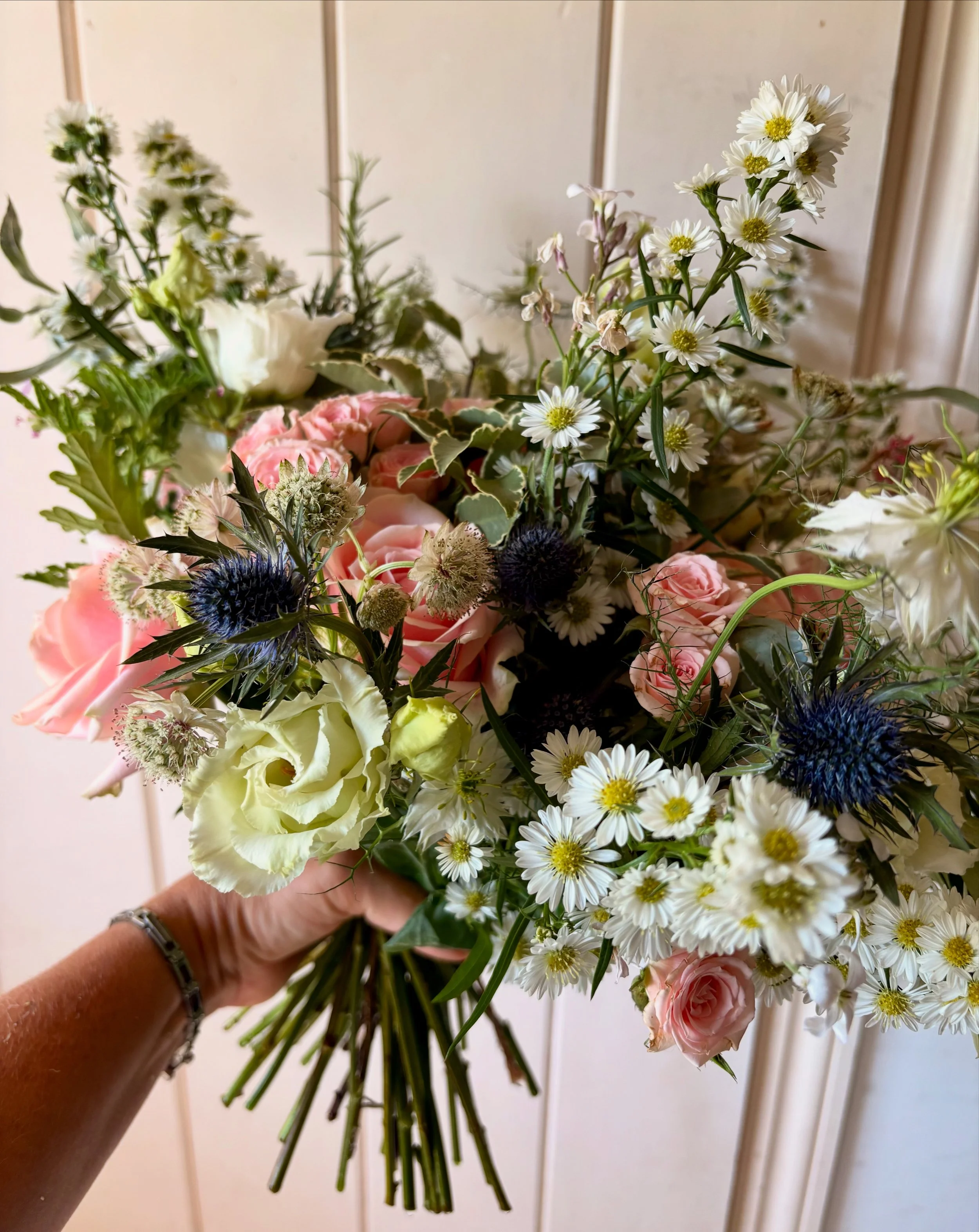 A hand holding a colorful bouquet of various fresh flowers, including pink roses, white lisianthus, purple thistle, and white daisies, against a neutral background.