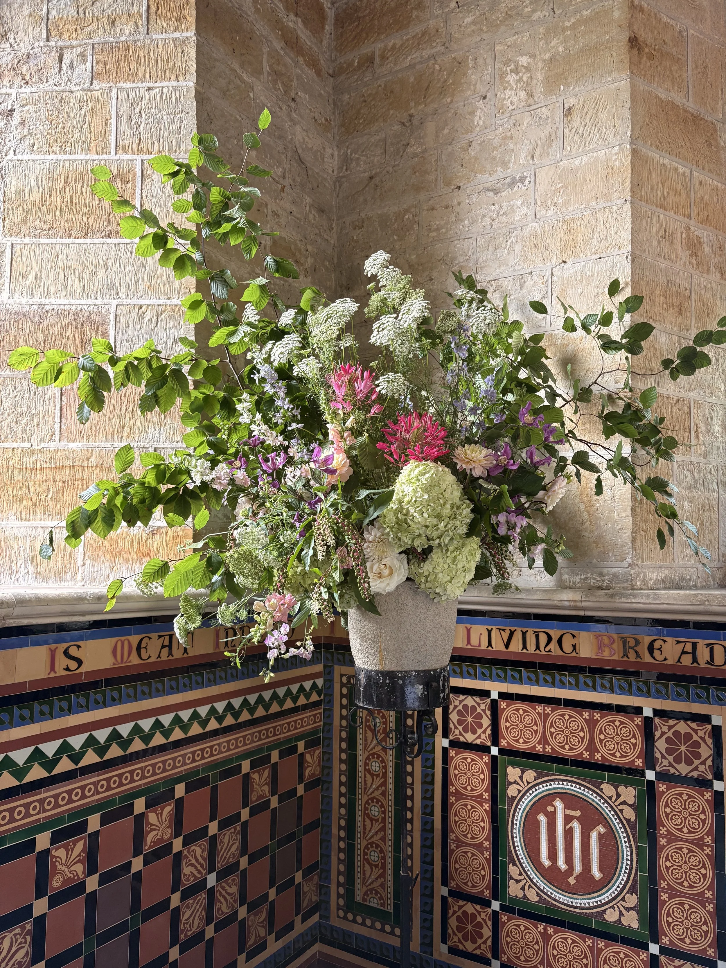 A tall floral arrangement in a white vase with a black stripe near the bottom, placed on a decorative tiled surface against a brick wall. The arrangement includes green leaves, white flowers, and pink, purple, and lavender flowers.