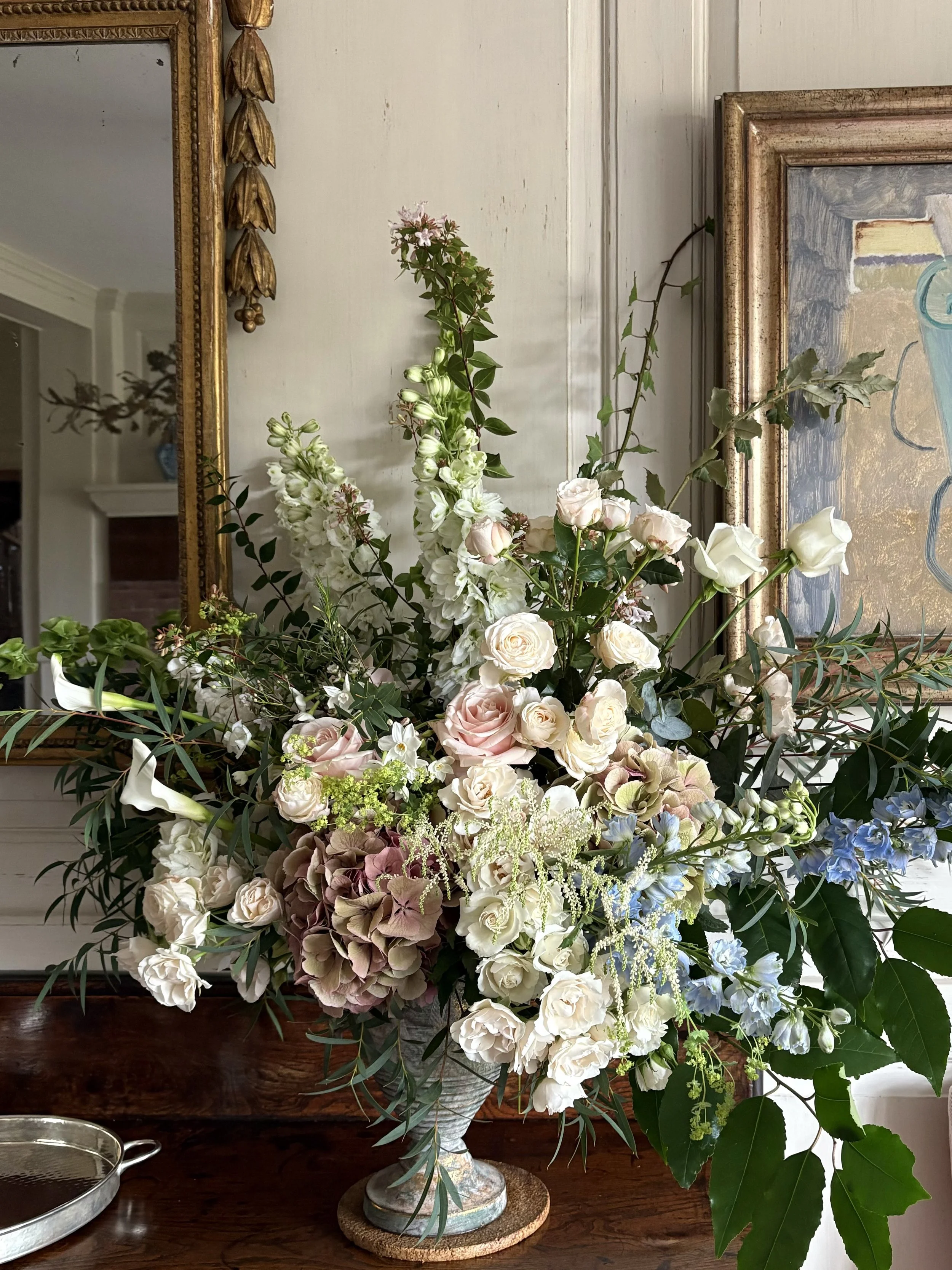A large floral arrangement with white, pink, and blue flowers, including roses, hydrangeas, and other greenery, in a decorative vase on a wooden surface.