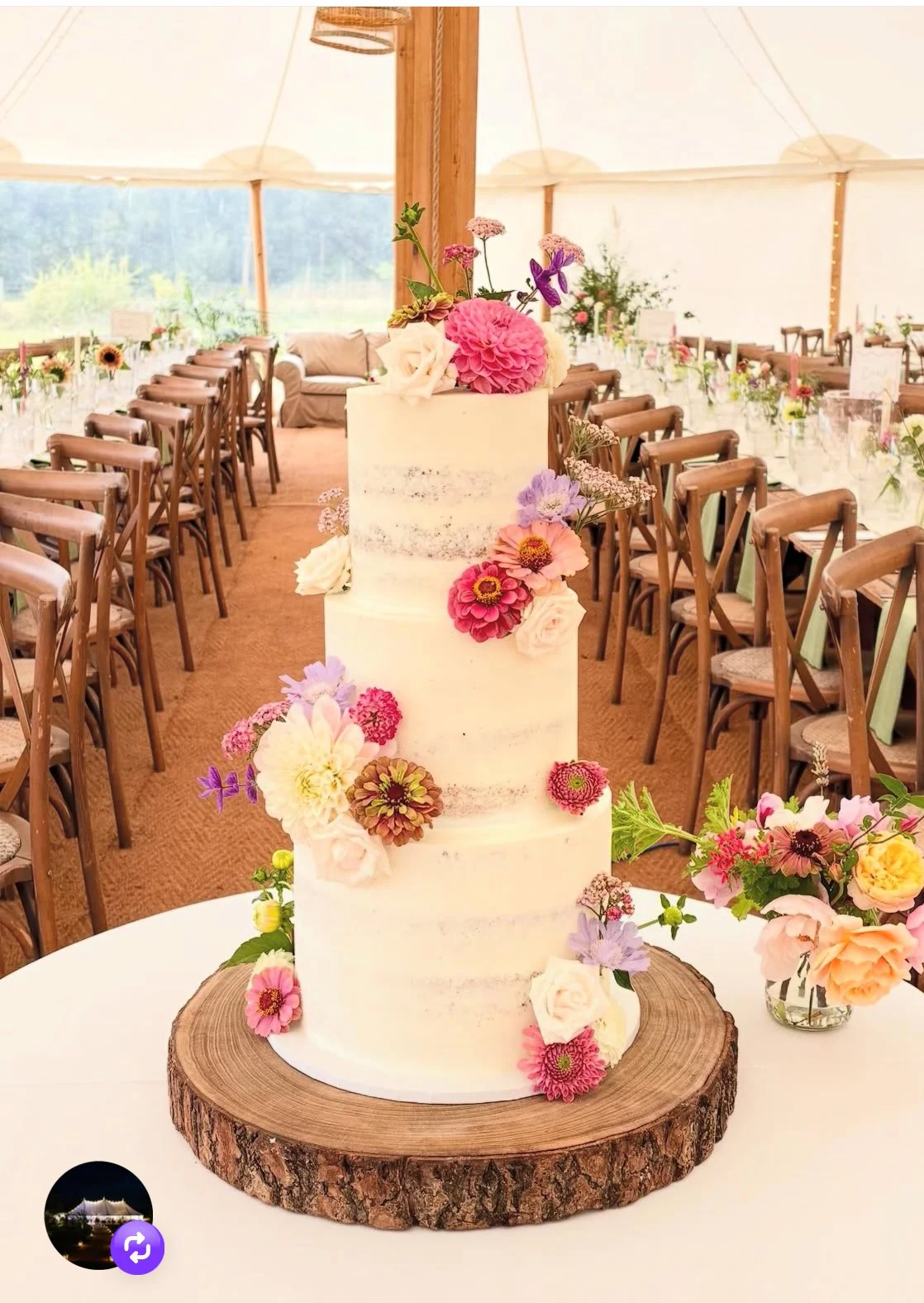 A four-tiered white wedding cake decorated with colorful flowers, placed on a wooden cake stand, inside a decorated tent for a wedding reception.