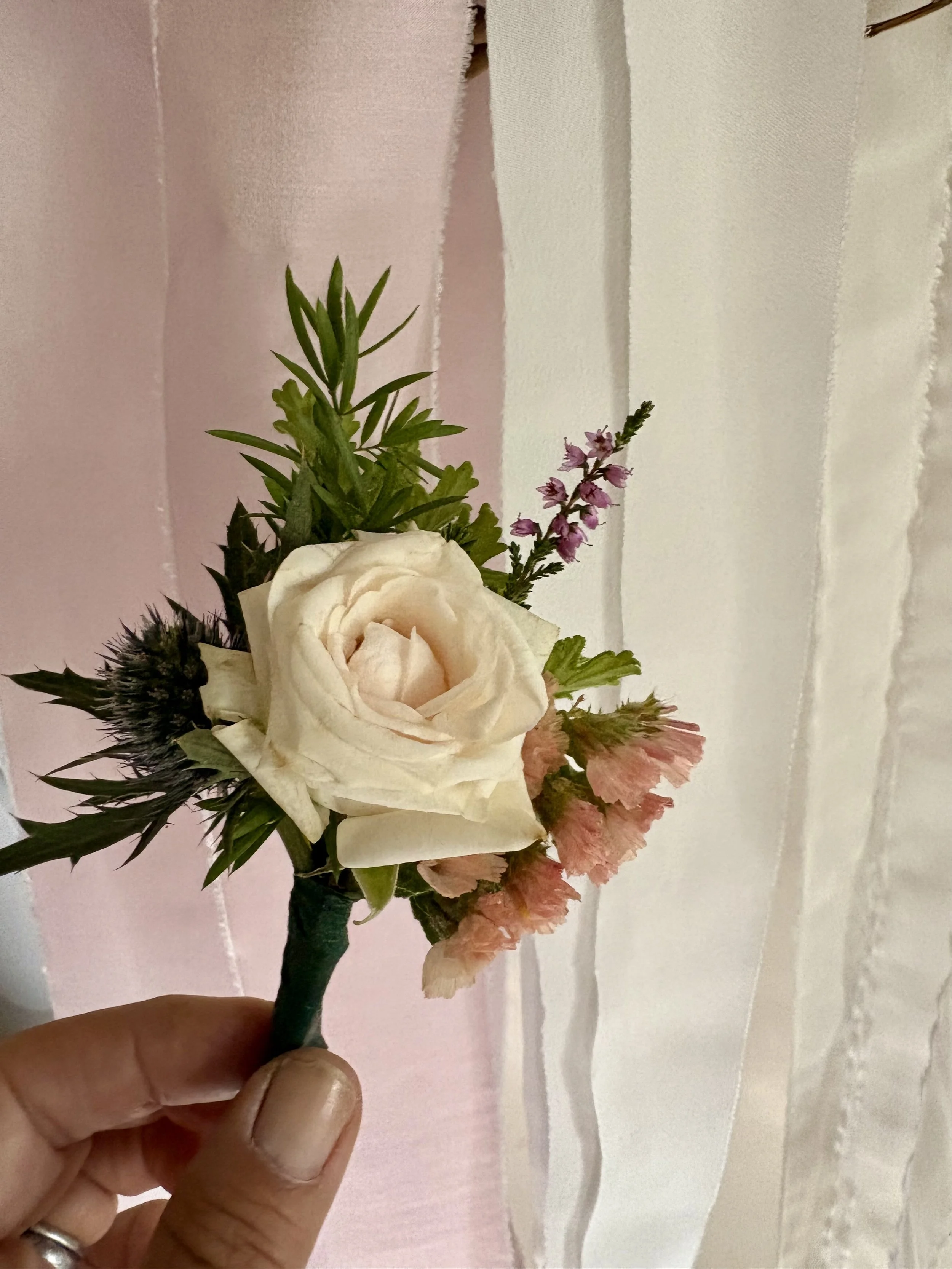 A hand holding a small bouquet of artificial flowers including a white rose, pink flowers, purple flowers, and green foliage against a pastel pink and white background.