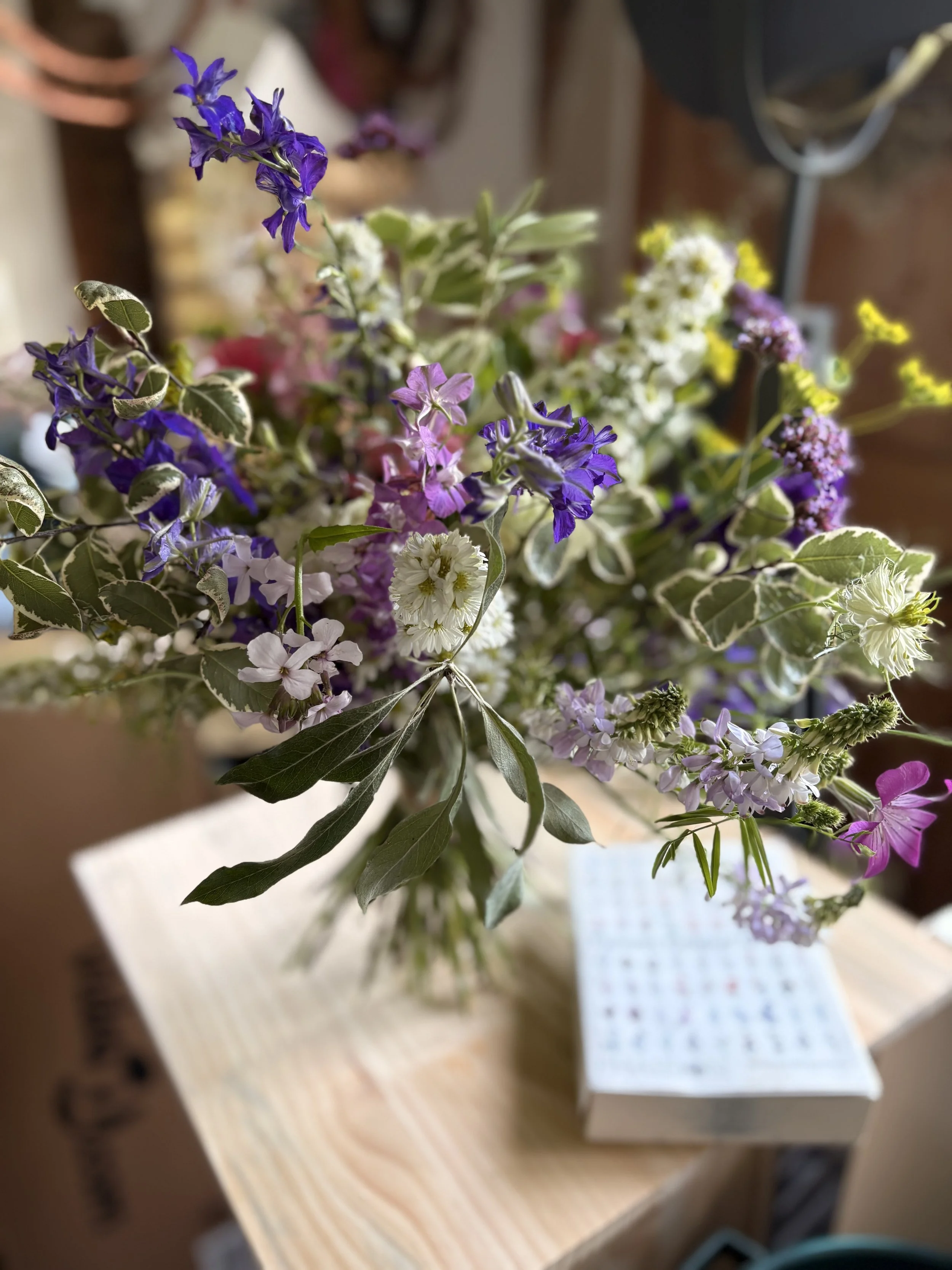 A bouquet of mixed flowers including purple, white, and pink blooms on a wooden table, with a blurry background.