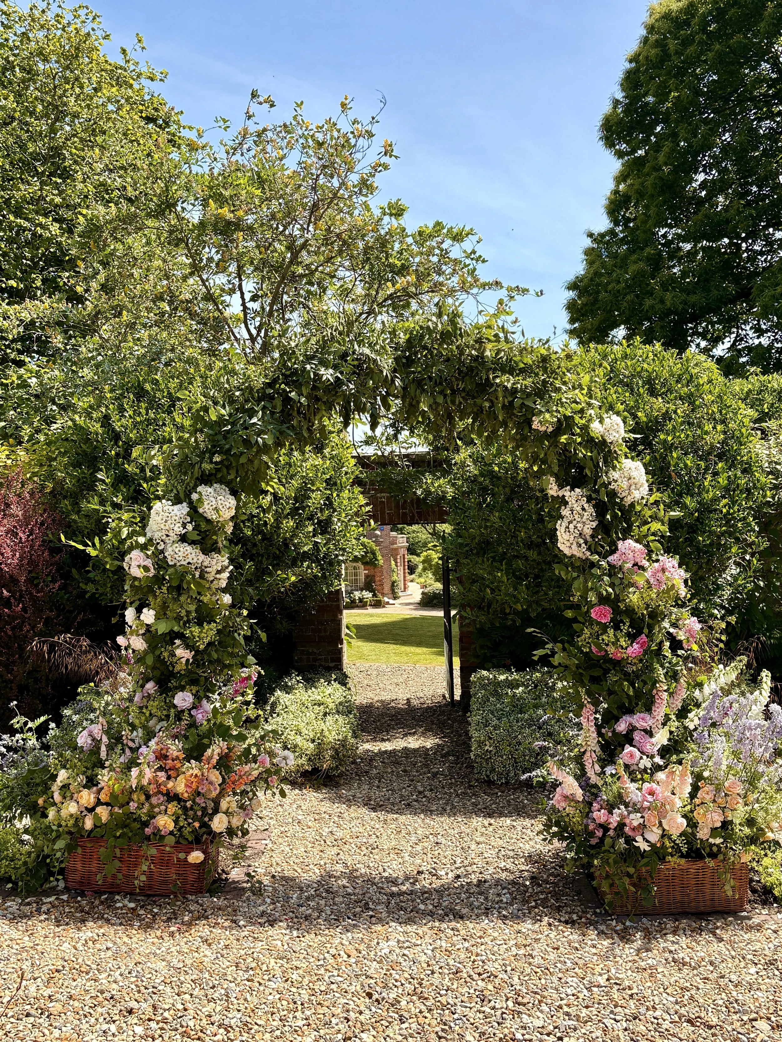 A floral archway made of greenery and pink and white flowers over a gravel pathway leading to a garden with a house in the background.